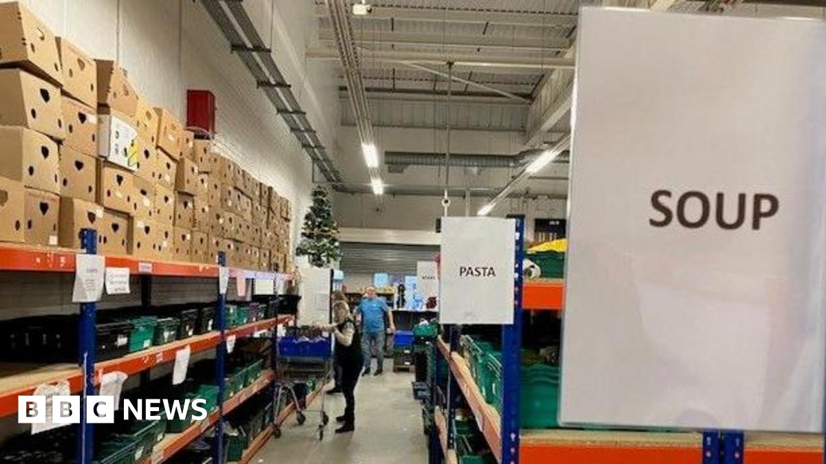 A long aisle in a warehouse stacked with boxes and cans of food. There are signs saying "Pasta" and "Soup". A woman with a trolley is in the distance picking up cans.