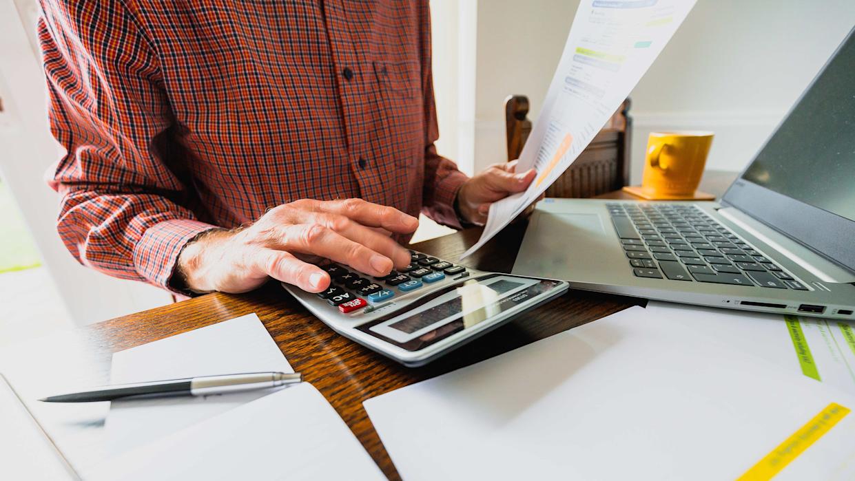 Wide angle image depicting a senior man's hand holding an energy bill while the other hand checks the numbers on a calculator. The table is strewn with documents and also a laptop. Room for copy space.