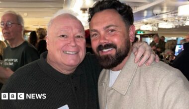 Michael Starke, 68, who as a bald head and wears a dark brown jumper, smiles and poses with his arm around the shoulders of Ray Quinn, who has dark hair swept back and a dark beard and wears a beige woollen jacket.