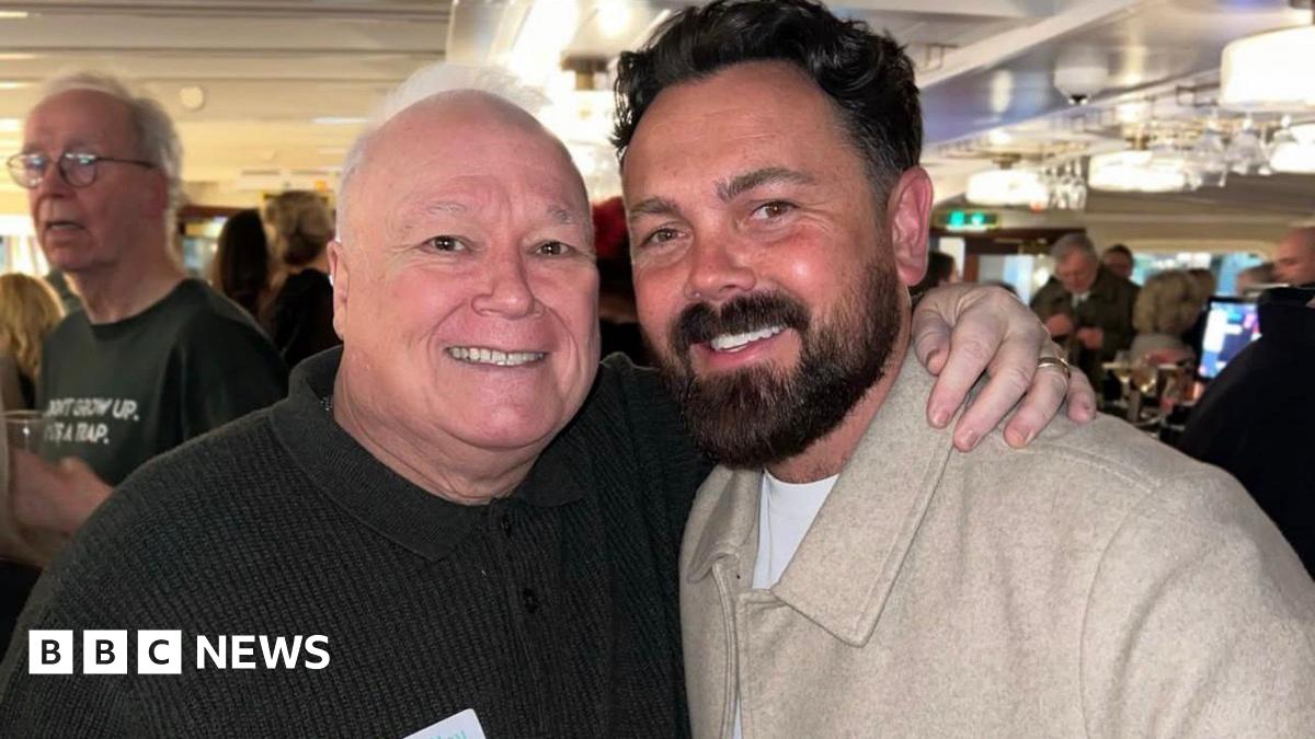 Michael Starke, 68, who as a bald head and wears a dark brown jumper, smiles and poses with his arm around the shoulders of Ray Quinn, who has dark hair swept back and a dark beard and wears a beige woollen jacket.