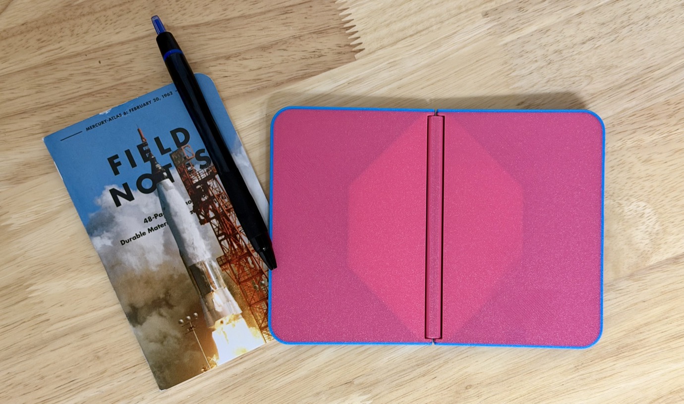 A Field Notes notebook with a pink cover and blue edges lies open on a wooden surface next to a black pen with a blue tip and a magazine featuring a rocket launch.