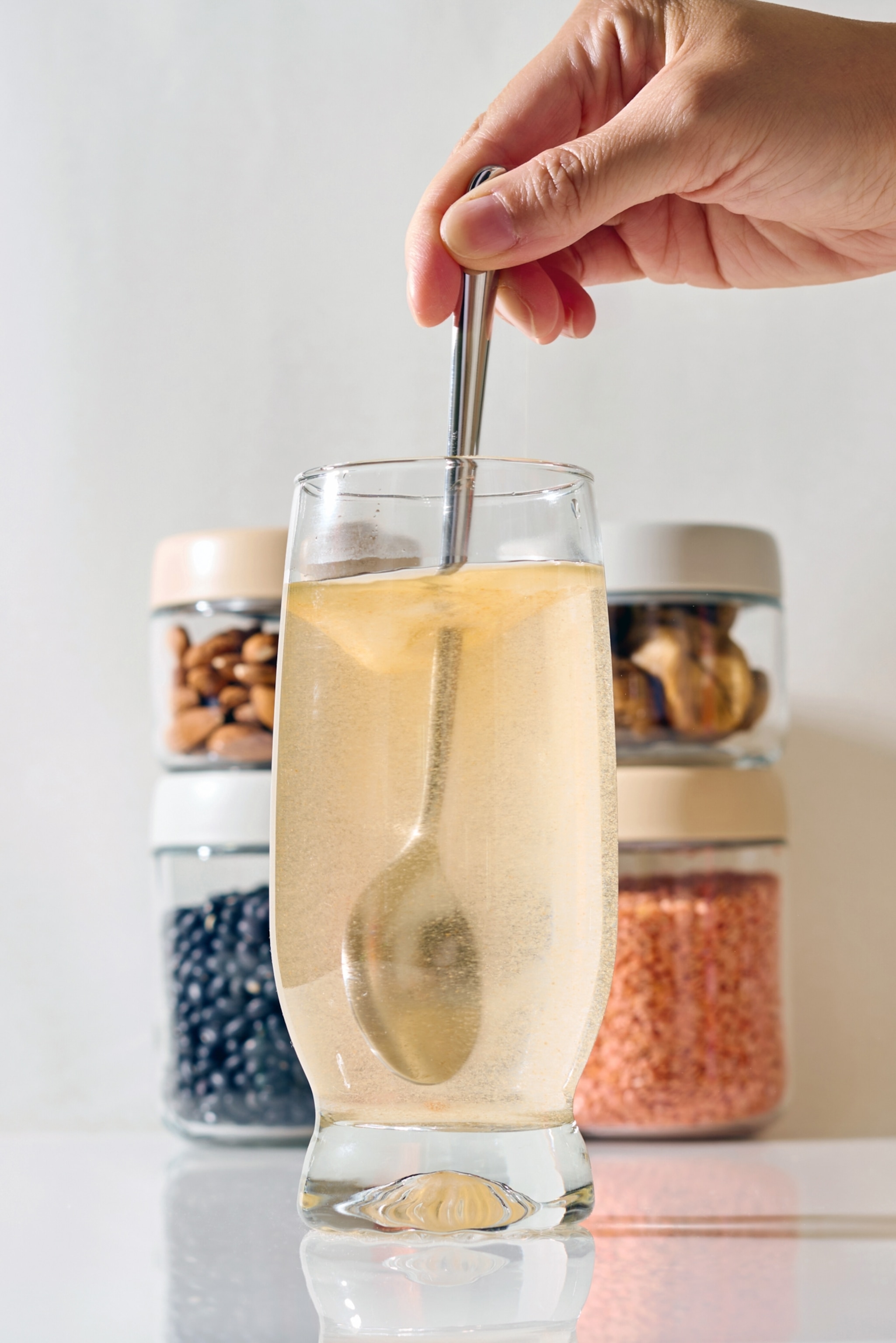 A hand stirs some fiber supplement in a glass of water with a metal spoon, set against jars of nuts and grains.