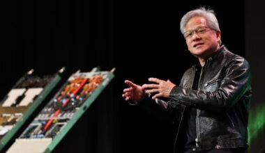 Jensen Huang gestures while speaking, seated on stage with computer hardware components displayed nearby.