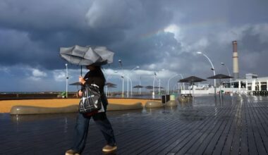 A person walks on a wet boardwalk with a windblown umbrella during stormy weather in Tel Aviv, with dark clouds and a faint rainbow overhead.