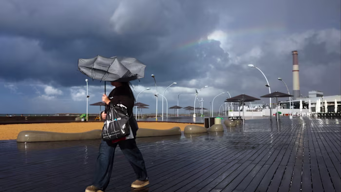 A person walks on a wet boardwalk with a windblown umbrella during stormy weather in Tel Aviv, with dark clouds and a faint rainbow overhead.