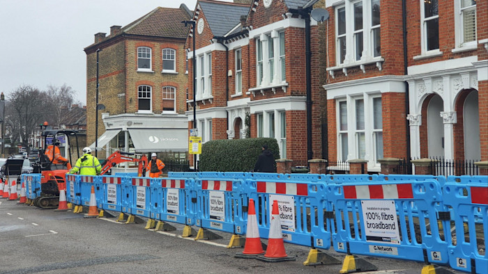 Workers in high-visibility clothing install fibre optic broadband behind blue barriers on a residential street in Clapham, London.