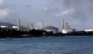 El Palito Refinery with large storage tanks and towers seen from the water, with palm trees lining the shore.