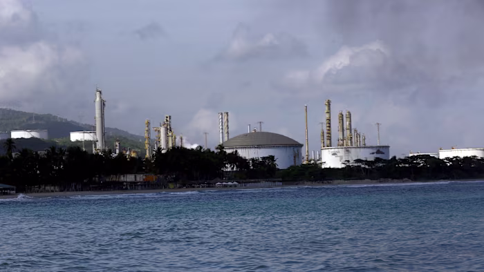 El Palito Refinery with large storage tanks and towers seen from the water, with palm trees lining the shore.