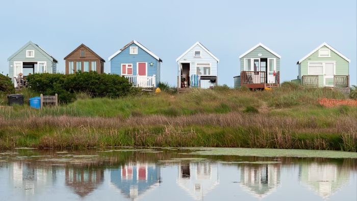 Five colourful beach huts in a row are reflected in still water at sunrise, with long shadows and empty beach grass in the foreground.