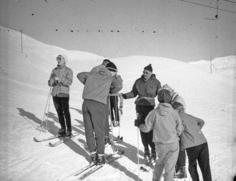 A group of people in winter clothing stand on a snowy ski slope, some holding ski poles. They appear to be preparing to ski, with snow-covered hills visible in the background. The image is in black and white.