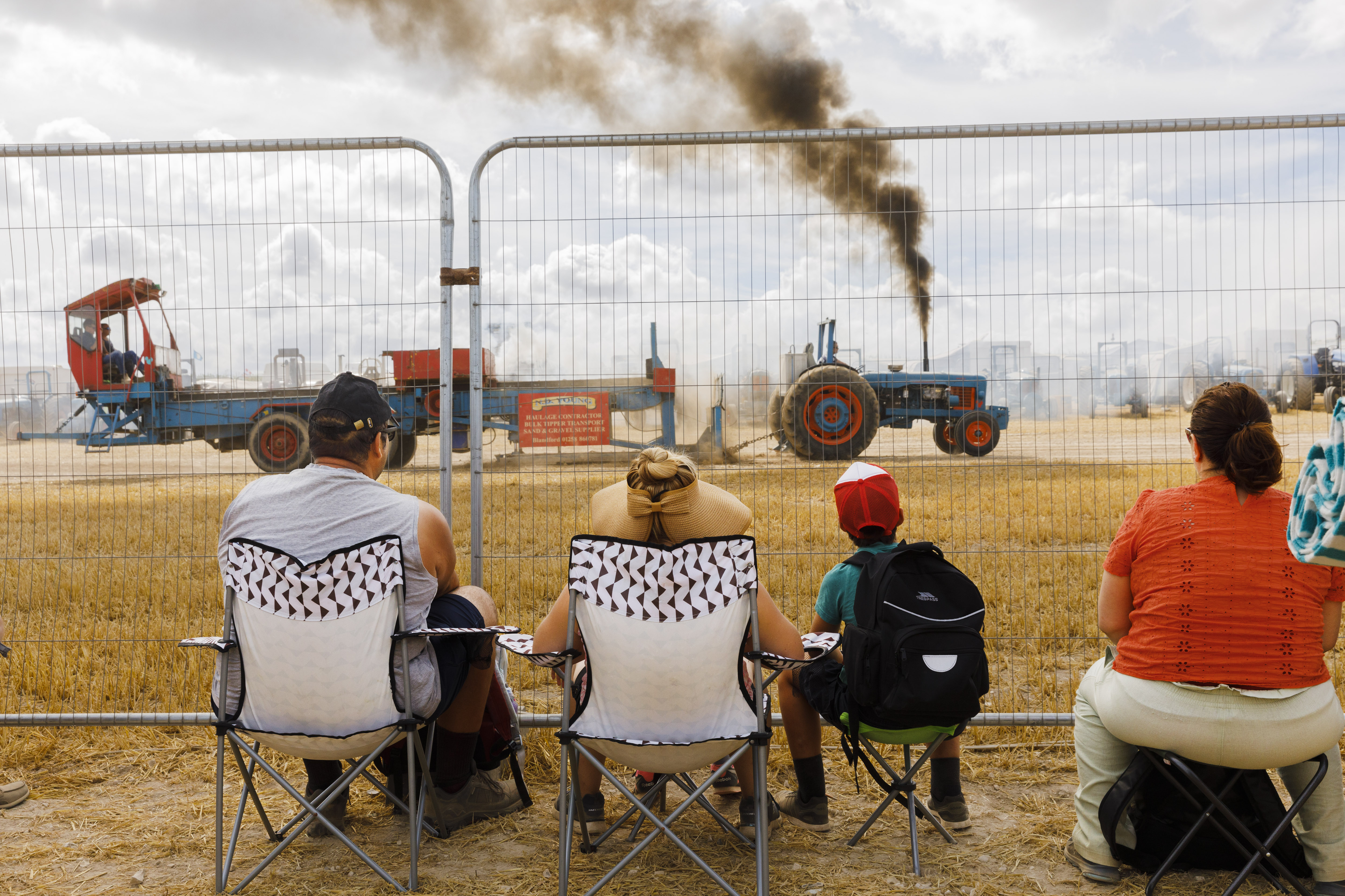 A family sits in folding chairs behind a wire fence, watching a blue vintage tractor emit a thick plume of black smoke during an outdoor exhibition.