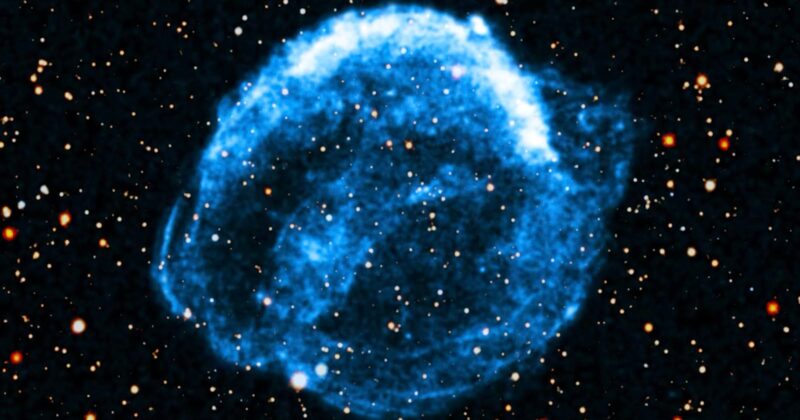 A glowing blue nebula appears as a circular, cloud-like formation against a dark background filled with scattered stars. The nebula glows intensely, with wispy edges and bright, varying light within it.