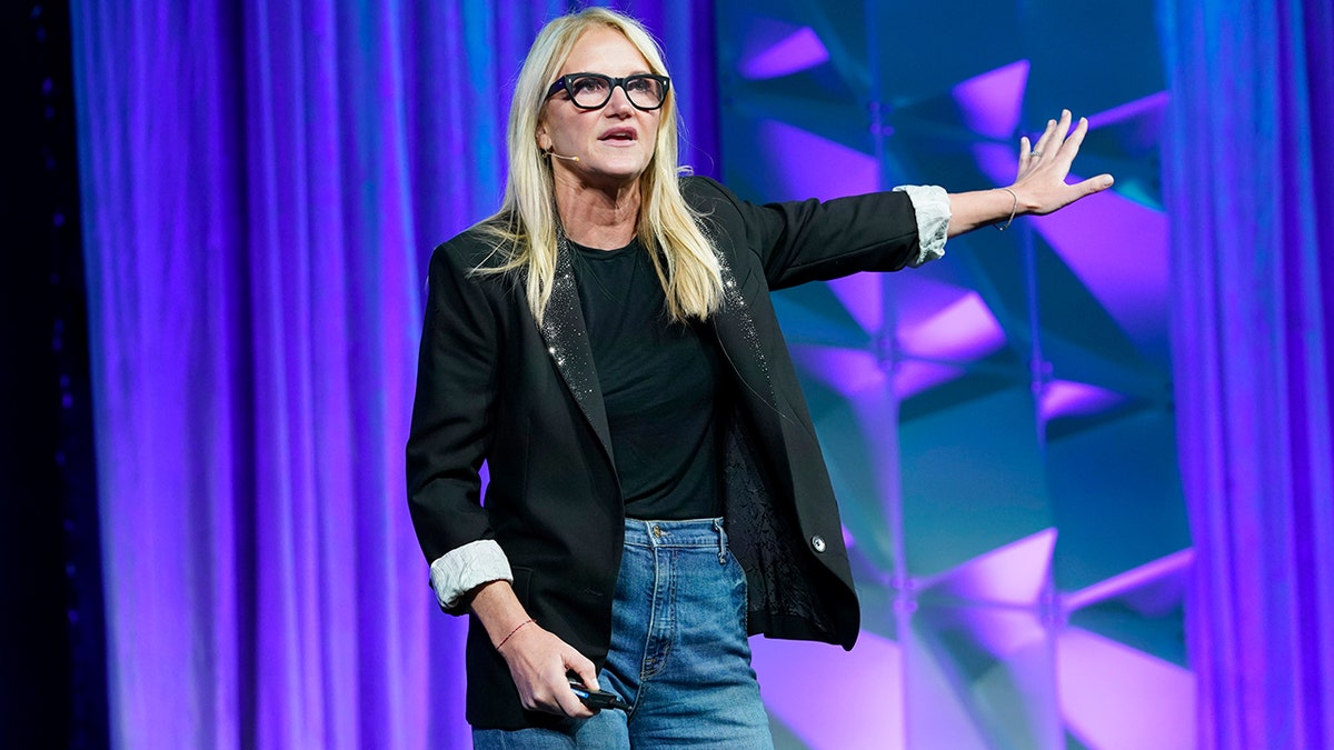 Mel Robbins on stage, wearing a black blazer and tee shirt, with blue jeans, holding her arm out to the side.
