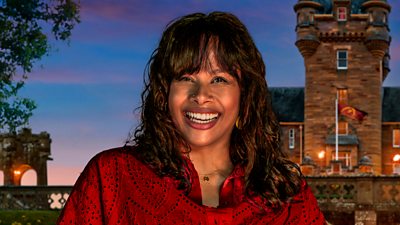 A woman in a red top smiles to camera, laughing, set against the backdrop of a castle by night
