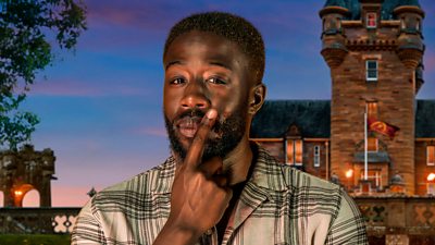 A man in a light shirt with a black plaid style pattern look to camera, holding a hand to his face in a gesture that suggests he's thinking about something. He's set against the backdrop of a castle by night.