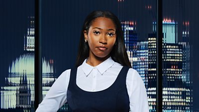 A woman in a shirt and pinafore dress poses outside the city backdrop at night