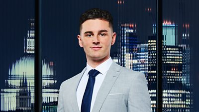 A man in a grey suit smiles outside a city backdrop at night 