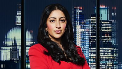 A woman (Nikki Jetha) in a red business suit stands outside a city landscape at night 