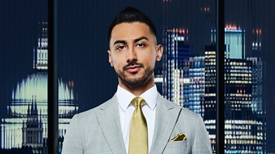 A man (Rajan Gill ) in a grey suit smiles outside a city landscape 