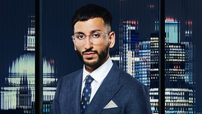 A man ( Tanmay Hingorani ) in glasses and a suit poses outside a city landscape 