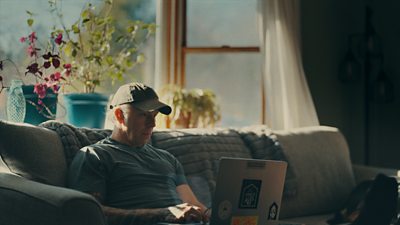 A man sits on a sofa in a living room and browses on a laptop. 