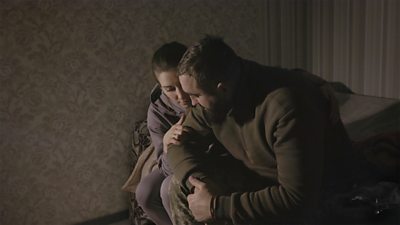 Two people embrace while sat on a sofa in a living room.