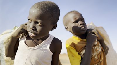 Two young children walk outside and carry heavy bags over their shoulders. 
