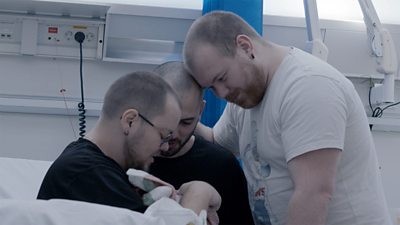 Three men gather around a hospital bed. One sits on the edge of the bed and holds a small bundle of blankets in his arms.