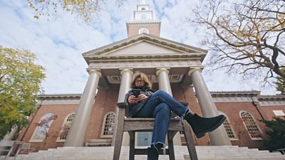 A woman sits in a wooden chair in front of a grand, building. 