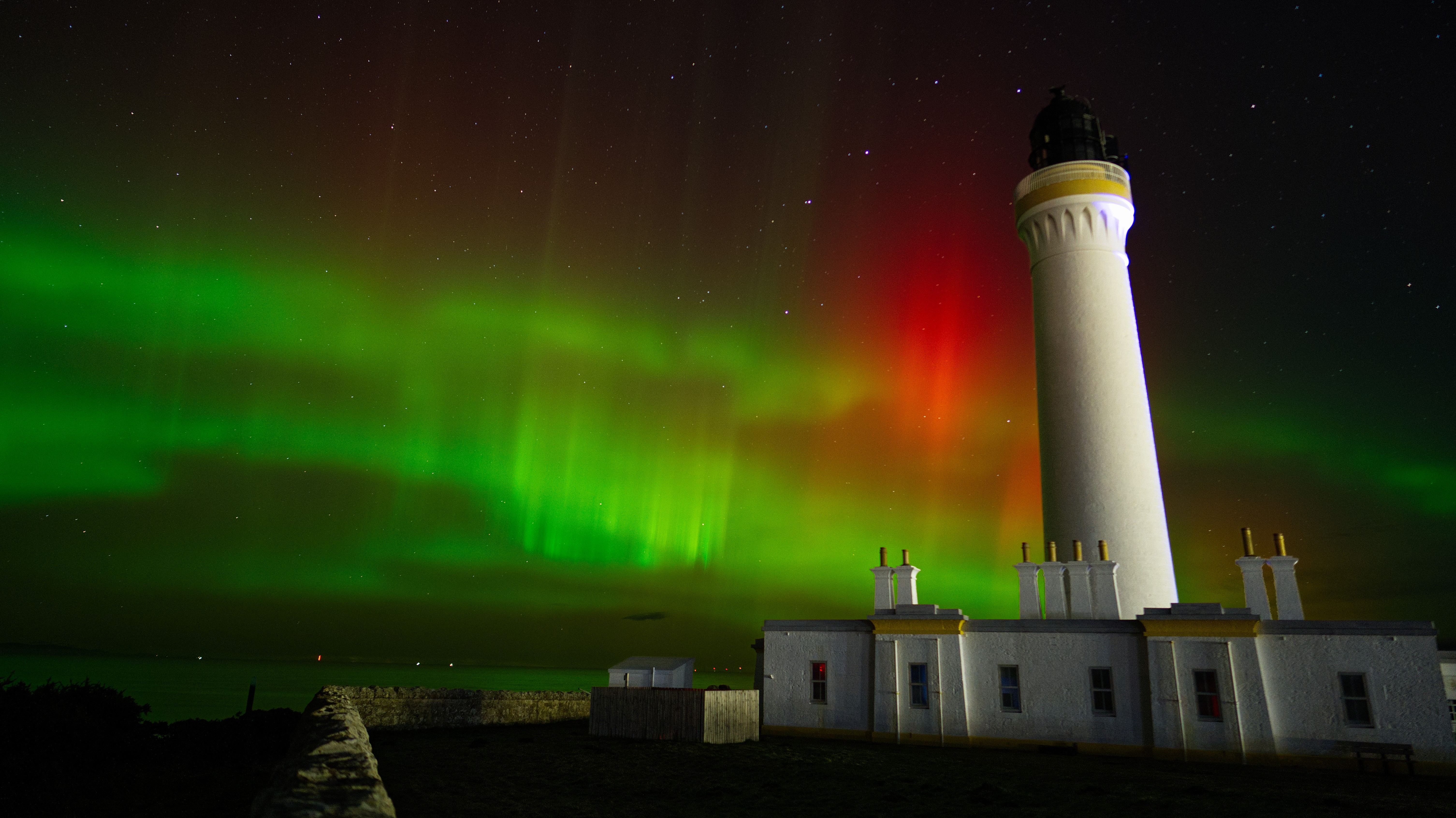 vivid green auroras and a patch of red in the sky behind a tall lighthouse structure.