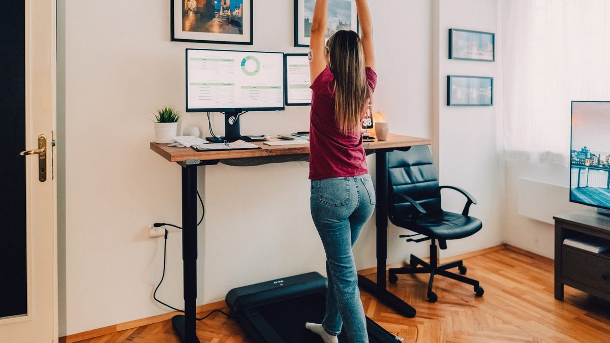 A person in jeans and a dark pink-purple tee using an under-desk treadmill while working on the computer.