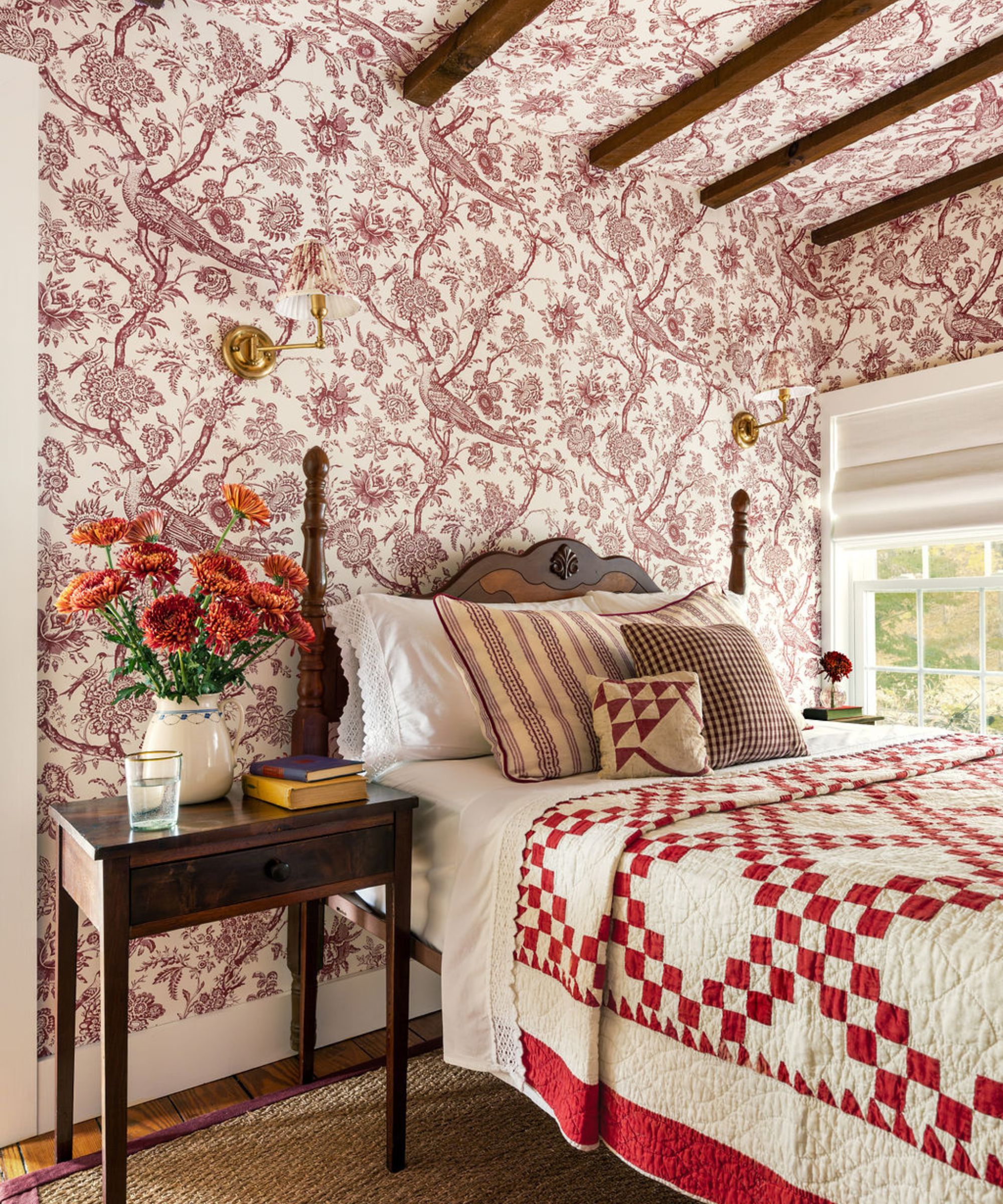 A cozy, traditional bedroom featuring burgundy floral toile wallpaper that extends onto the ceiling. The bed is dressed with a red and white checkered quilt and mixed pattern pillows, flanked by a dark wood nightstand with orange flowers and brass wall sconces