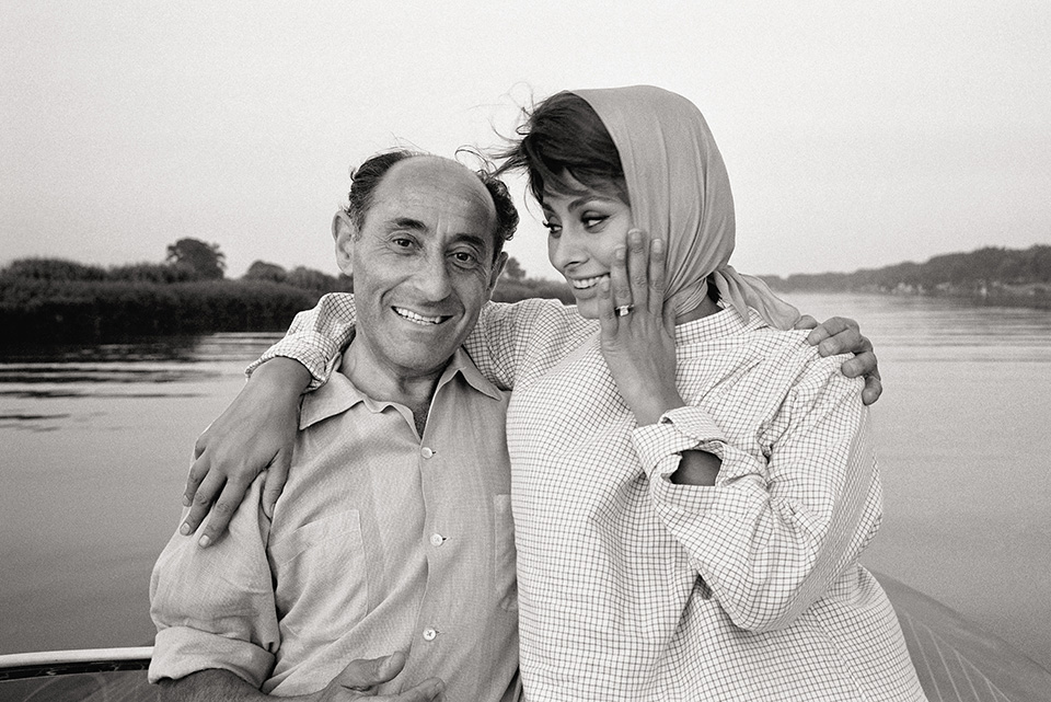 A candid black-and-white photo shows Sophia Loren wearing a headscarf and smiling as she rests her arm around a man&rsquo;s shoulder on a boat.