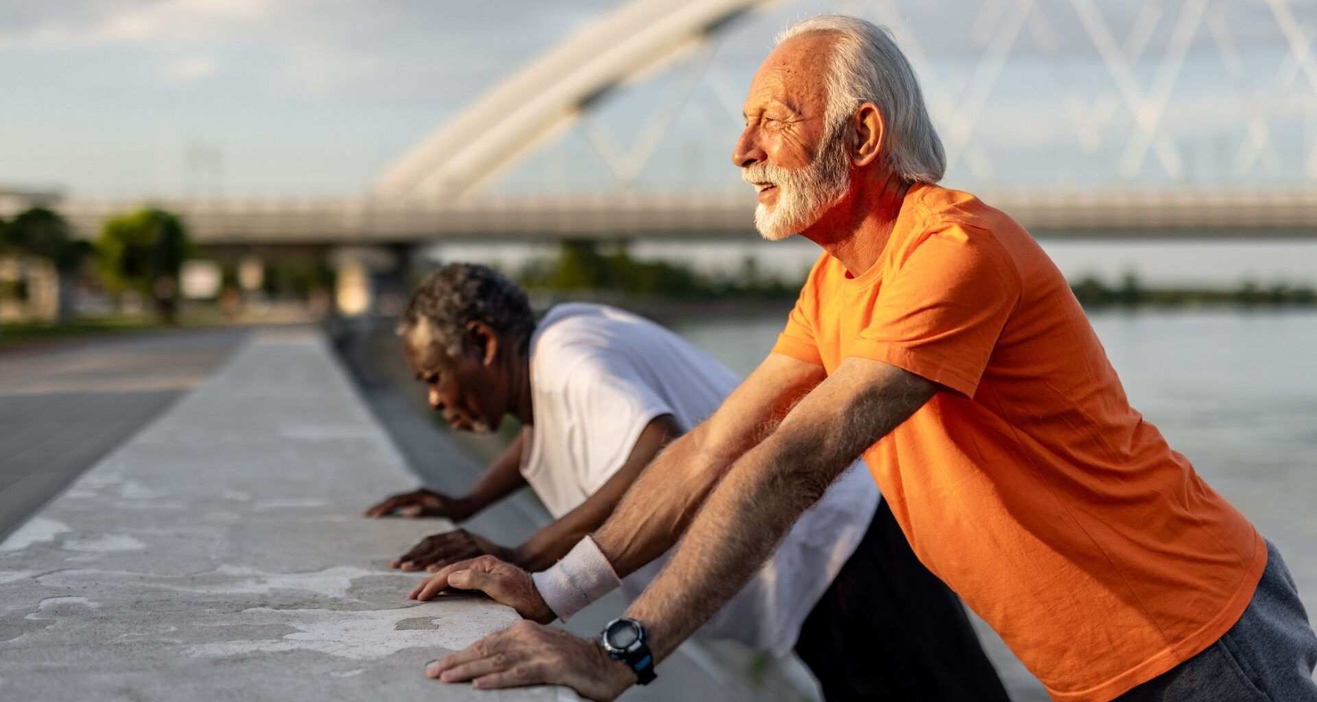 man in an orange tshirt and man in a white tshirt behind him both sideways to the camera performing press ups against a low wall outside.
