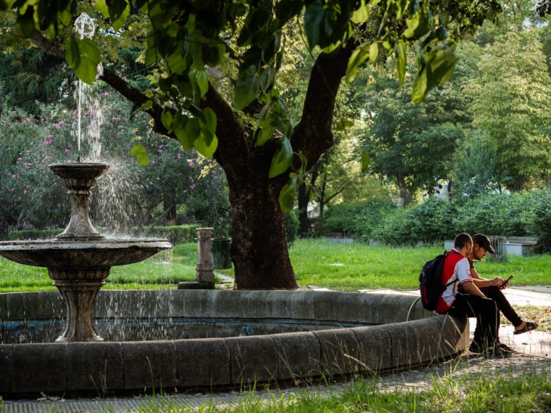 Two people sit on the edge of a circular stone fountain in a lush, green park, shaded by large trees. Sunlight filters through the leaves, and water splashes from the fountain's top tier.