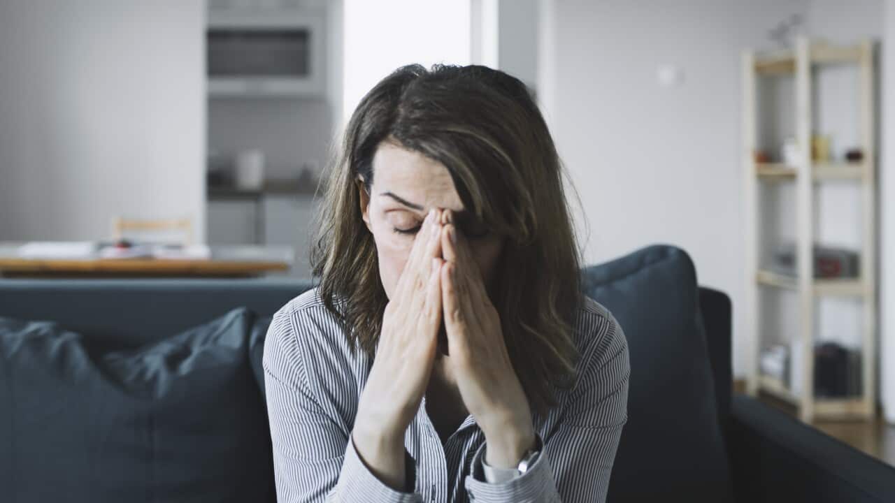 A woman is sitting on a couch with her hands on her face. She has a look of distress on her face.