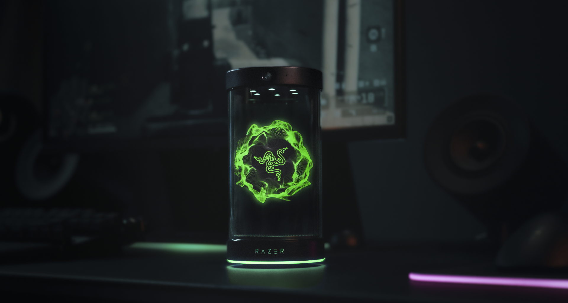 The Project AVA desk companion from Razer sits atop a black desk. A holographic image of Razer's logo lights up the screen in green.