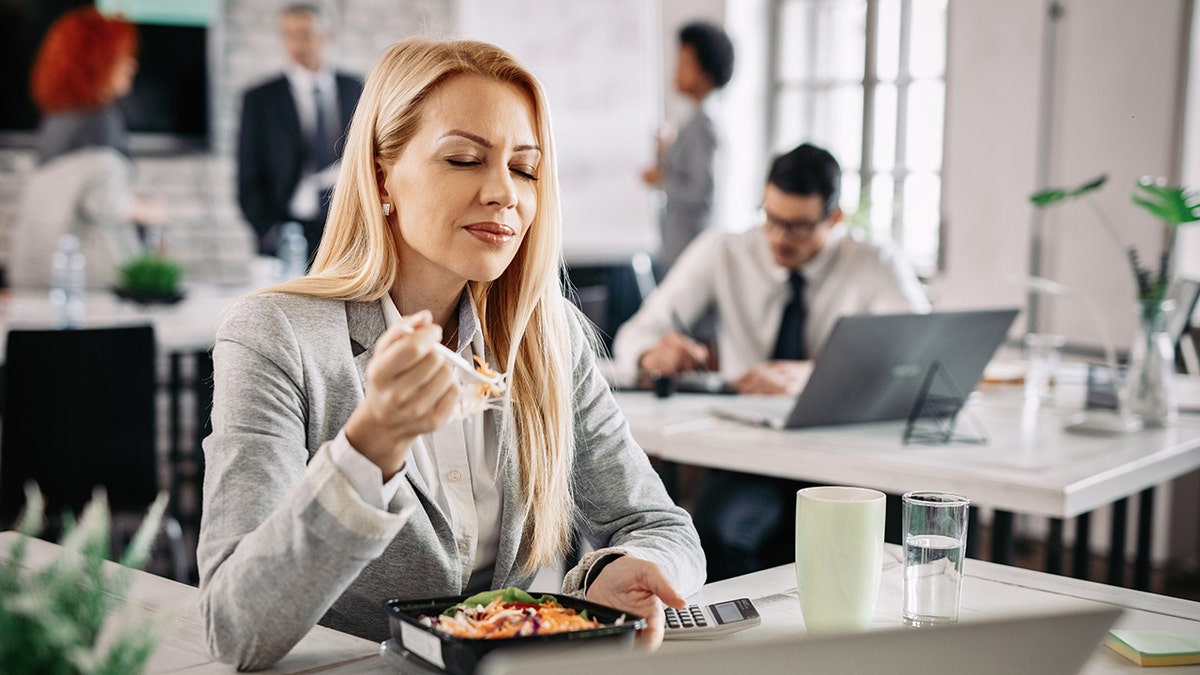 A woman enjoying healthy lunch, seen with eyes closed, in office breakroom or cafeteria, coworkers seen in background