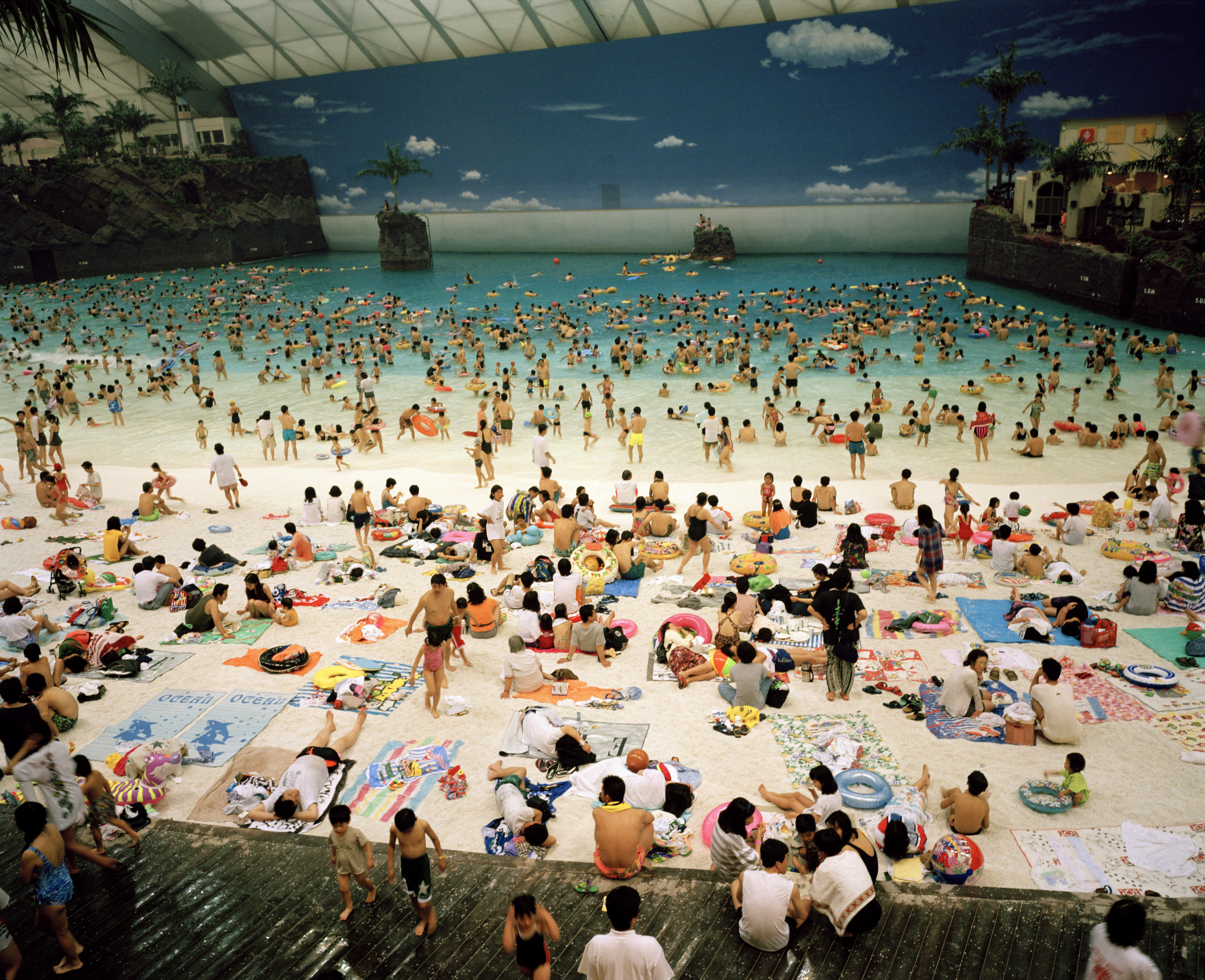 Thousands of people crowd a massive indoor wave pool and artificial beach under a giant dome featuring a painted blue sky with clouds.