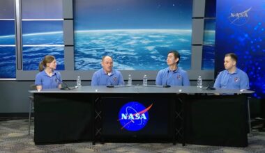 Four individuals wearing pale blue shirts sit behind a long table with a black cloth over it and the blue NASA circle logo with blue screens behind them.