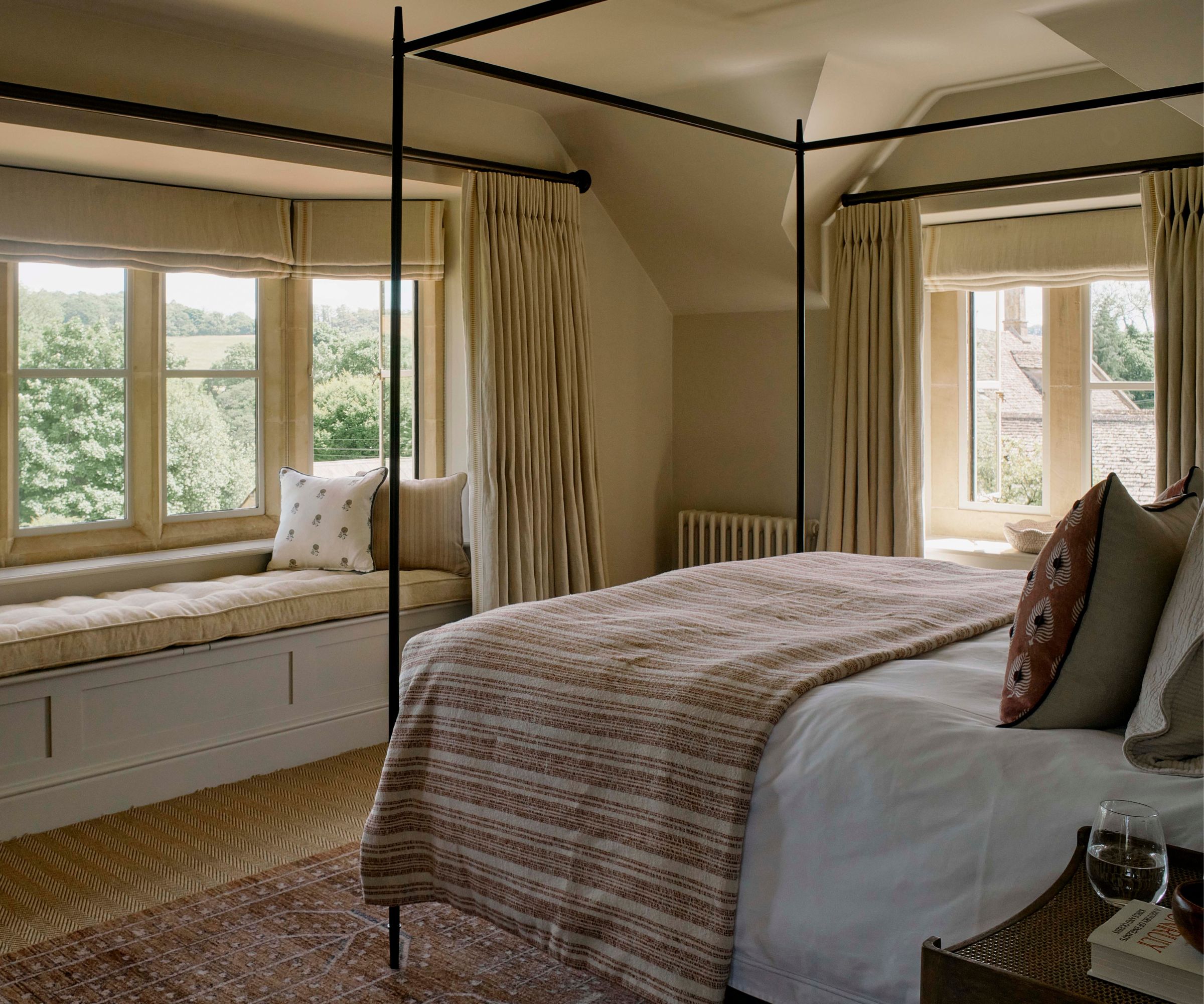 A warm neutral bedroom with a black four poster bed and a charming window seat