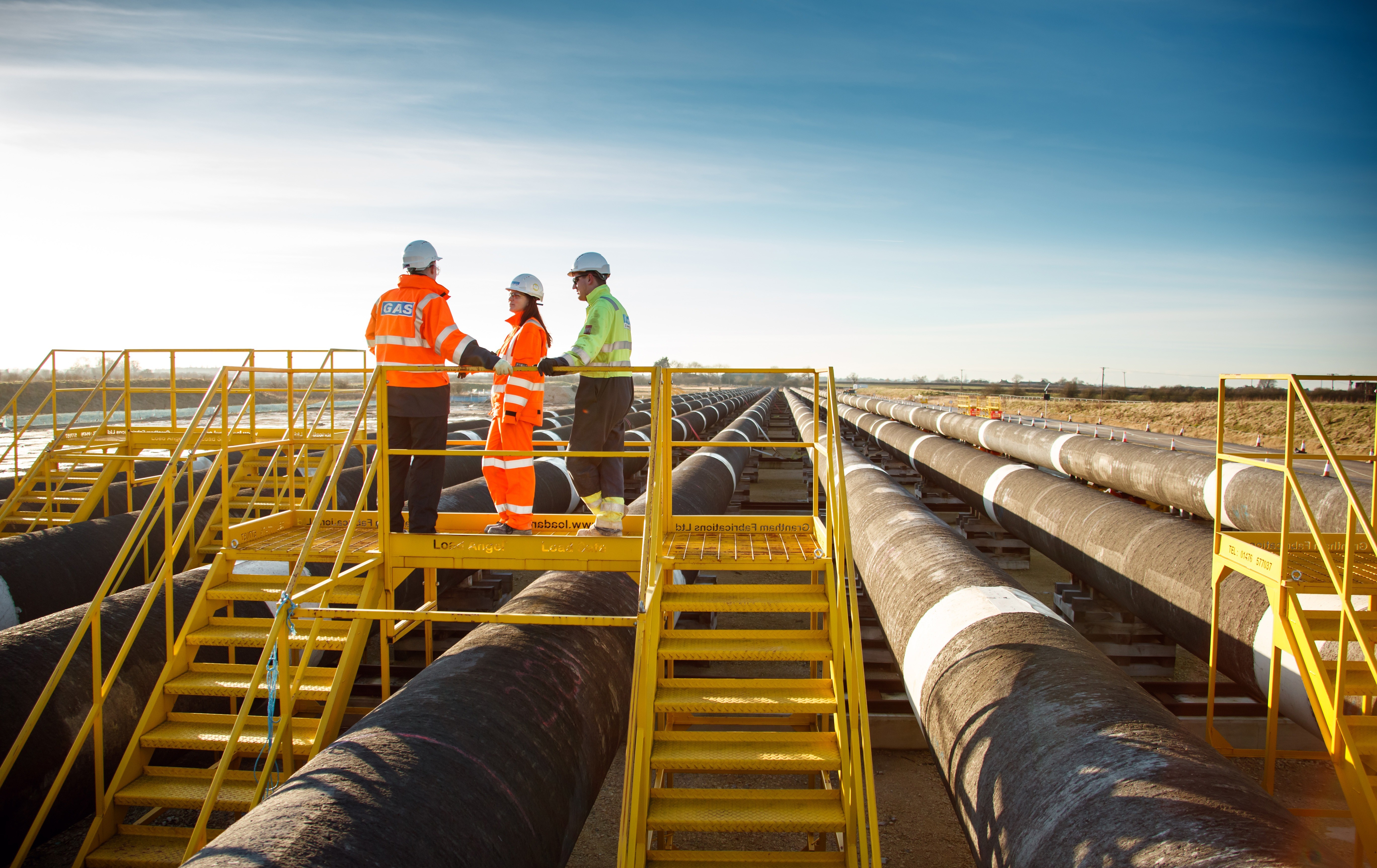 Three workers in safety gear stand on a yellow platform above gas pipelines, surveying the landscape.