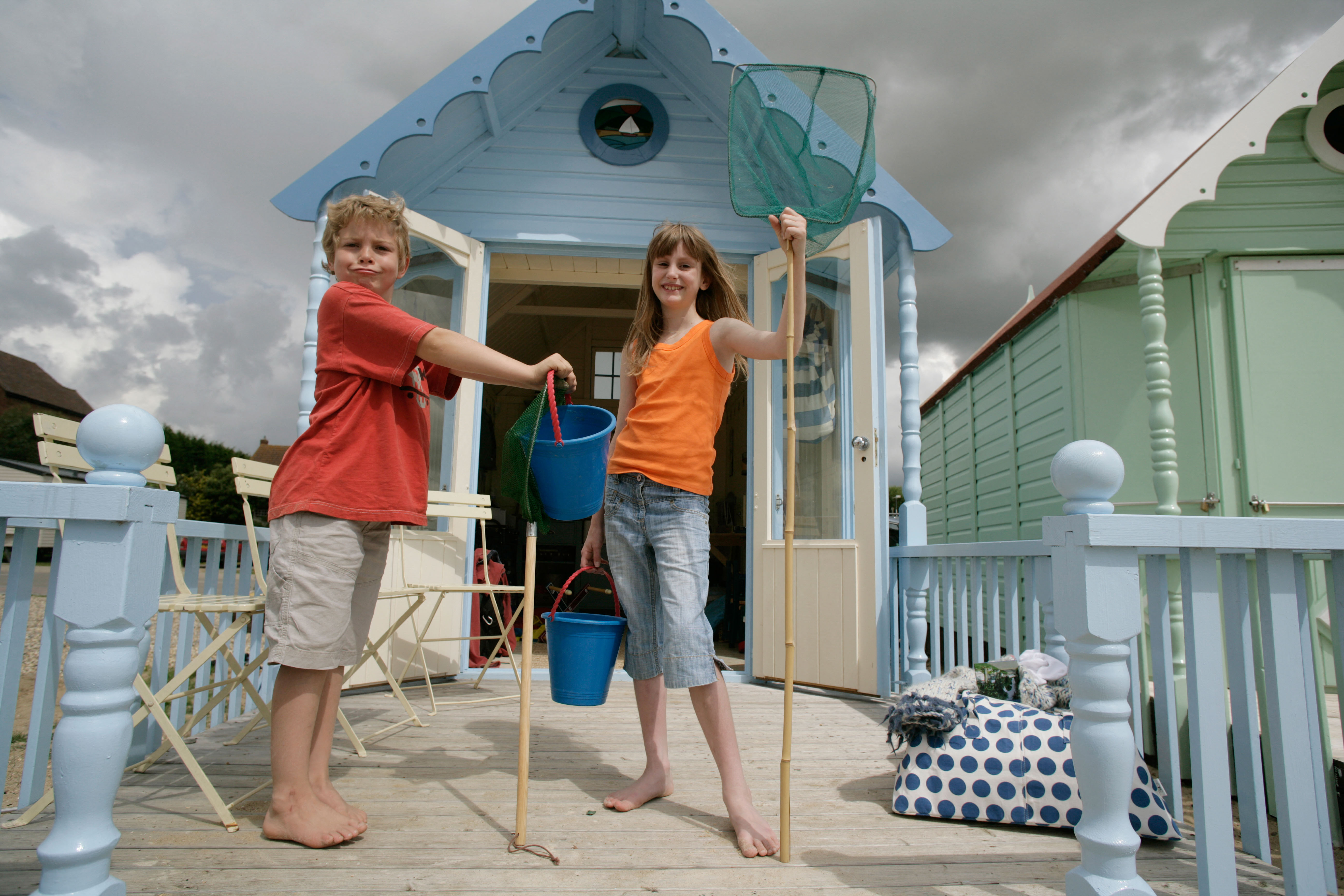 Two children standing barefoot on a wooden porch with fishing nets and buckets in front of a blue beach hut.