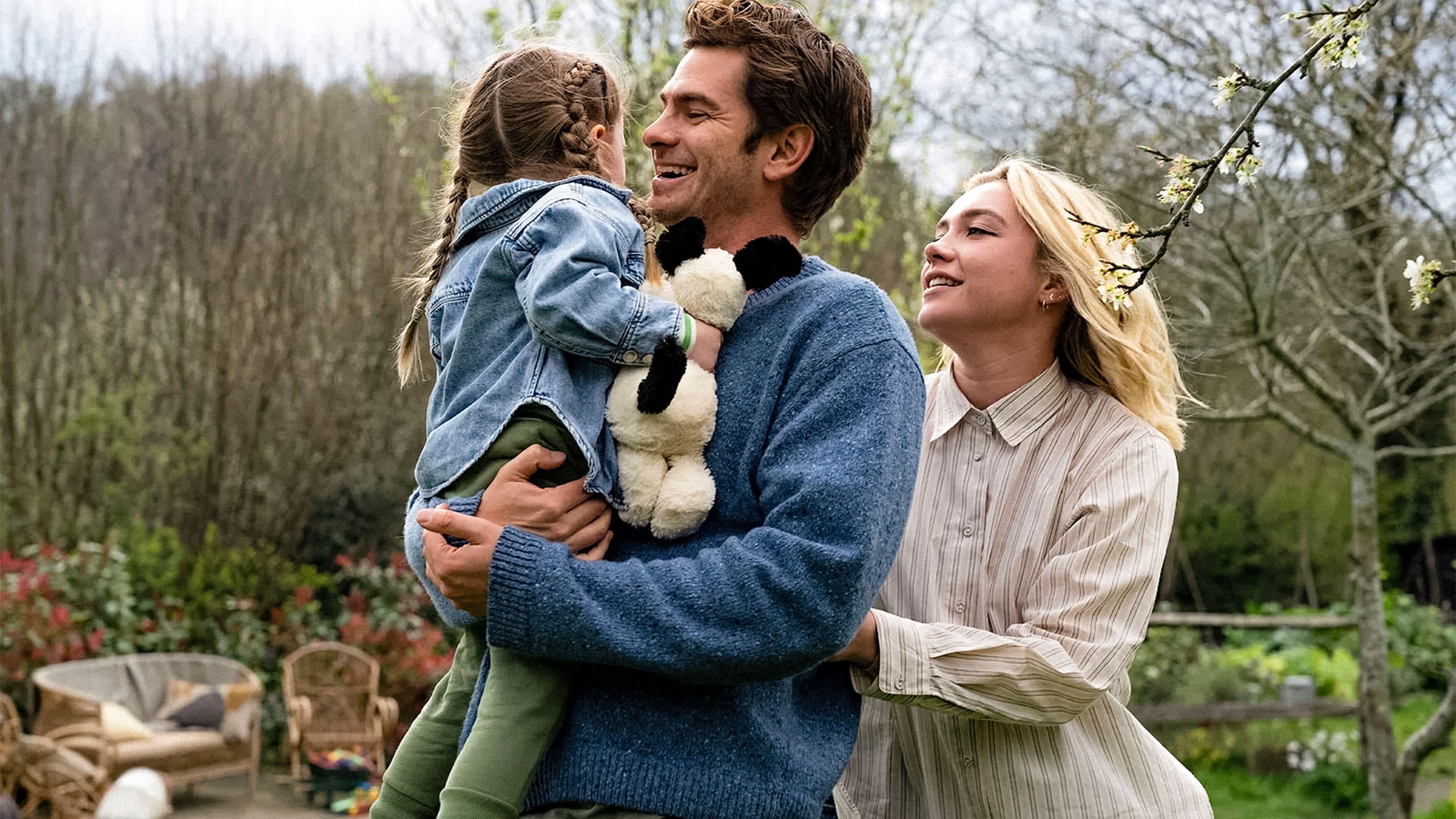Andrew Garfield holding a young child with pigtails and a panda toy, while Florence Pugh stands beside them, smiling.