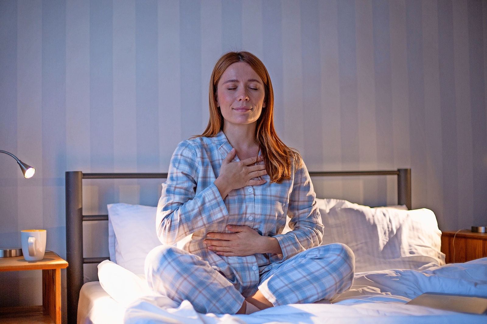 Woman in pajamas meditating in bed with her eyes closed, hands on her chest and belly.