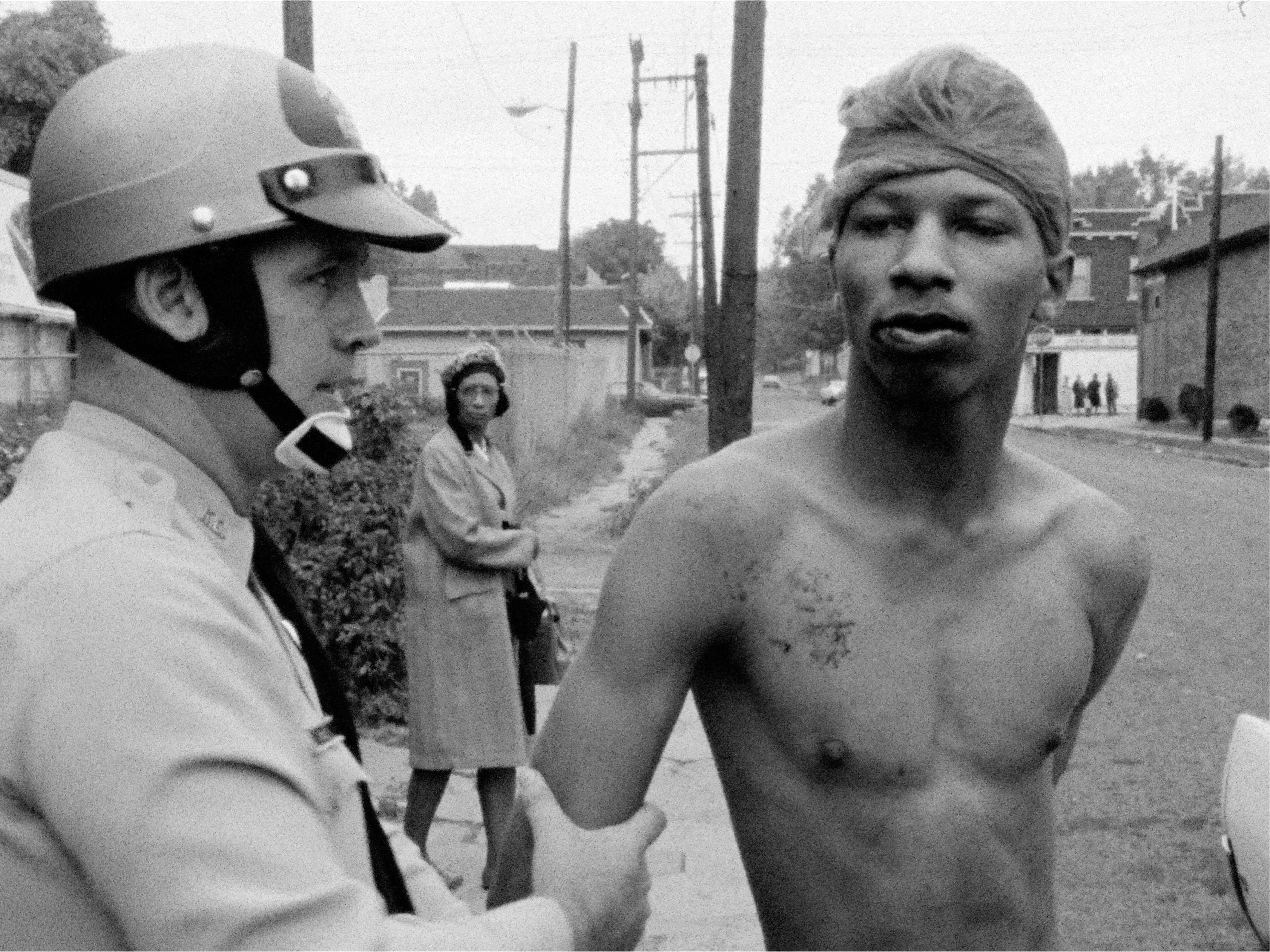 A police officer in a helmet holding the arm of a shirtless man wearing a head wrap, with a woman in a coat observing in the background.