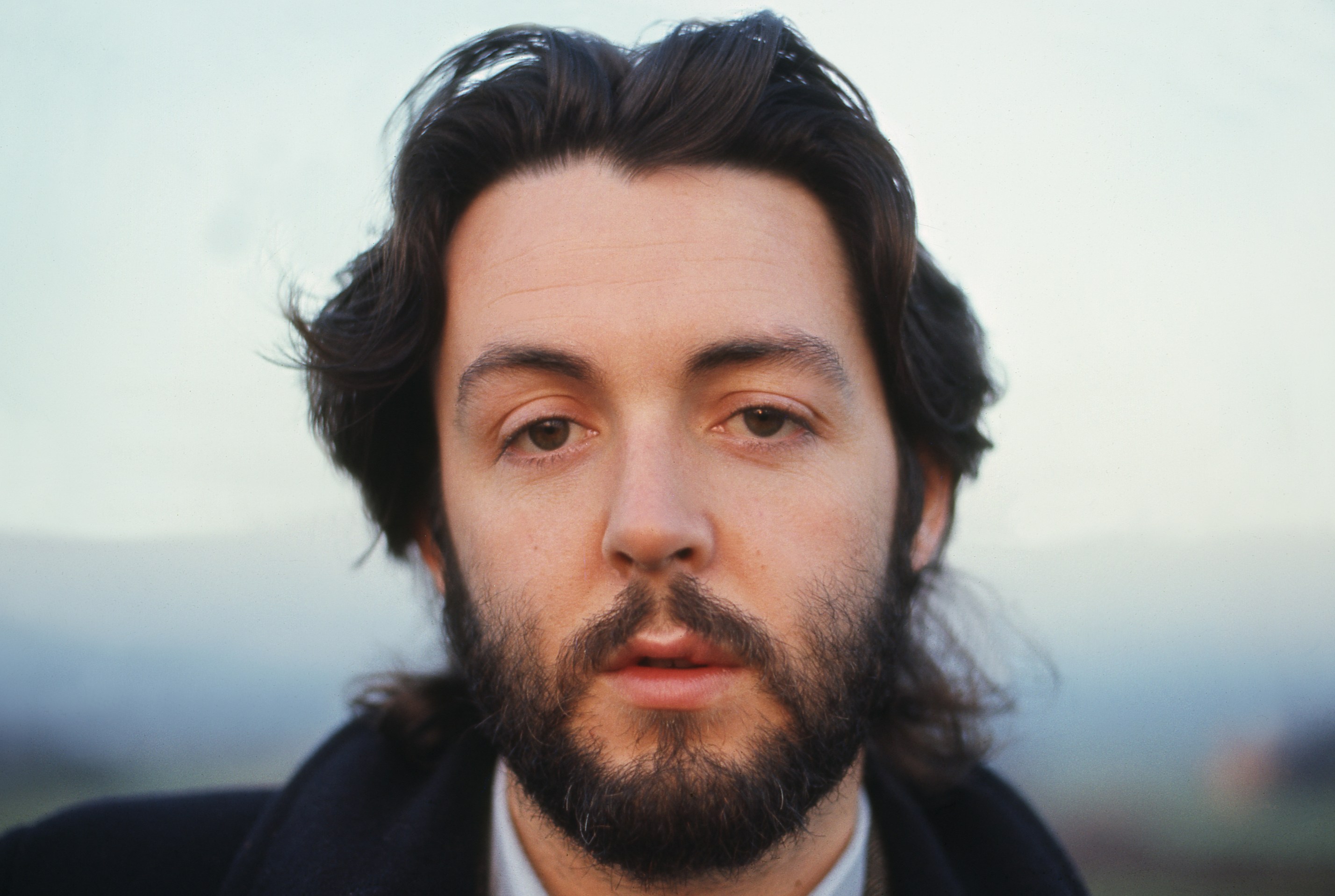 Paul McCartney in a close-up photo with long hair and a beard, looking directly at the viewer.