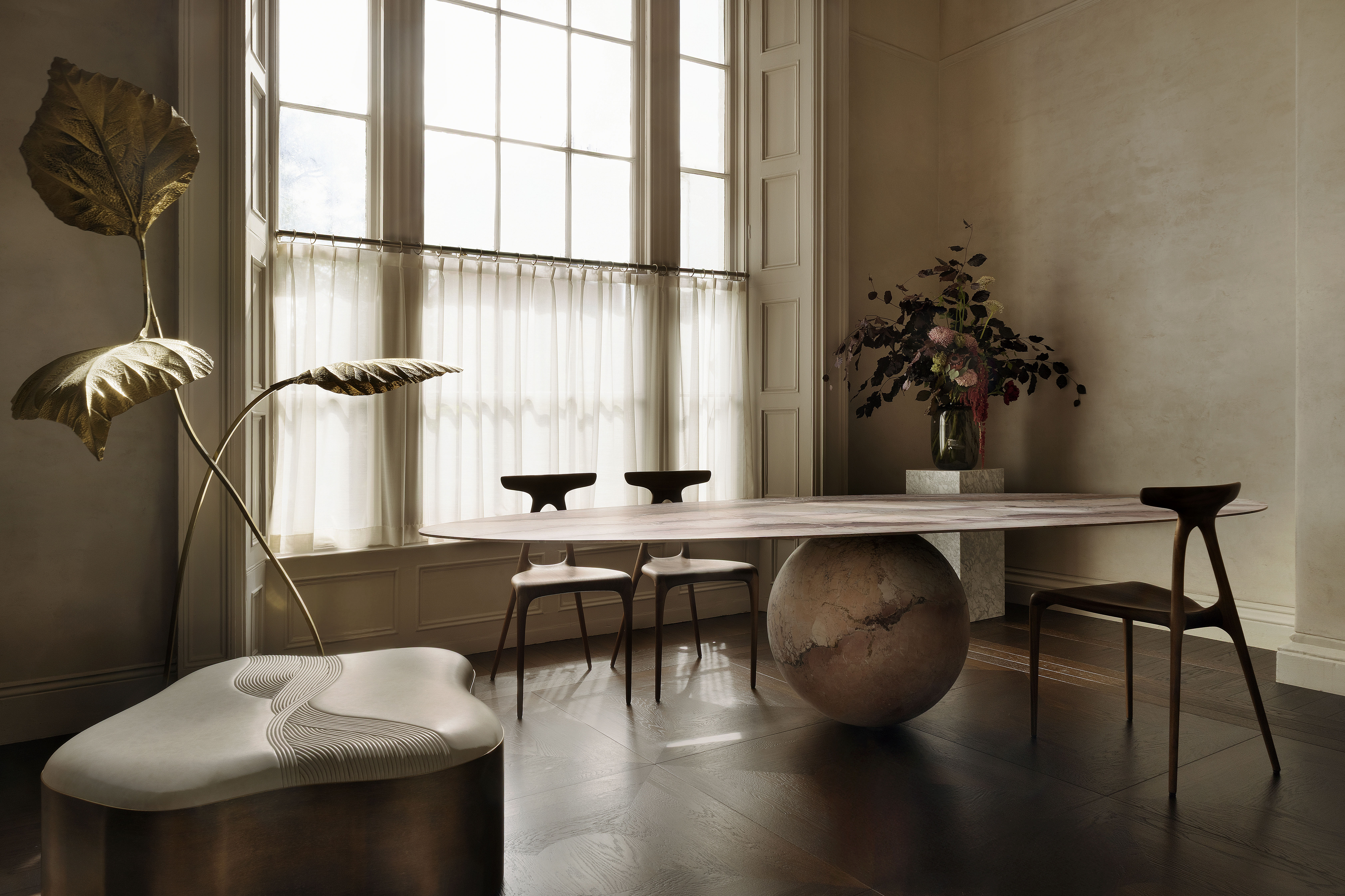 A dining room with a large window, a table with a spherical base and oval top, three chairs, an abstract light fixture with large gold leaves, and a low bench.