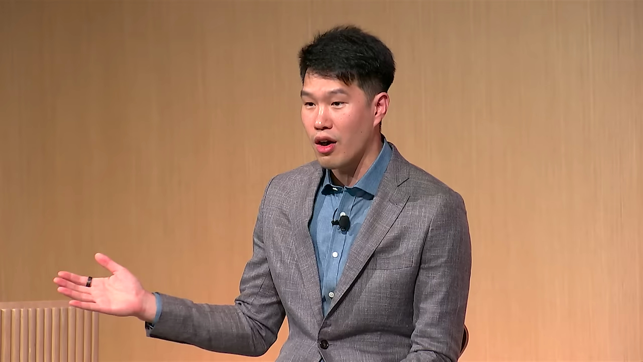 Mark Chen of OpenAI speaking during a fireside chat, gesturing with his left hand.