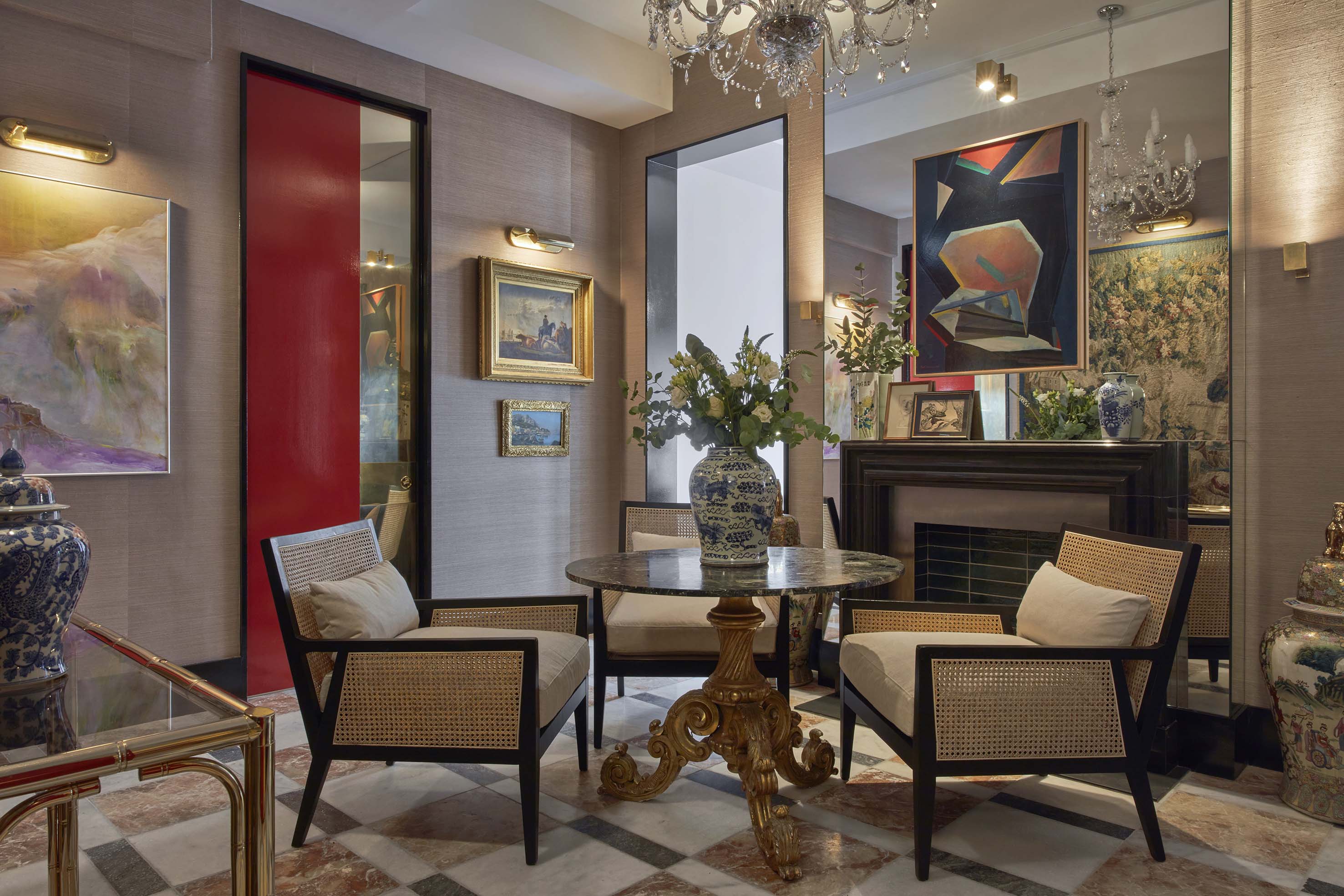 An ornate waiting room featuring a round marble table, two chairs with woven cane backs, a black fireplace, mirrors, and various artworks.
